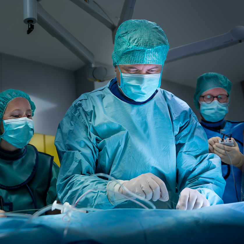 Prof. Dr. Benjamin Meder in surgical clothing leans over a patient. Other members of the surgical team can be seen in the background.