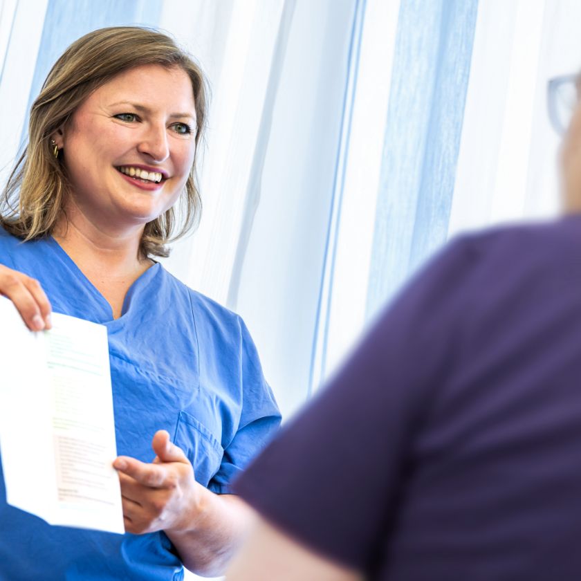 Nursing expert Lena Jung shows a patient an information sheet and smiles. The patient is blurred in the background.