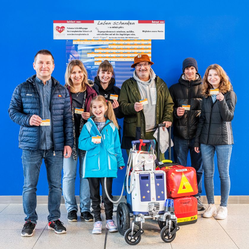 Group photo of six adults and Lara, the patient from paediatric heart surgery. Next to her is a trolley with the Berlin Heart. Everyone is looking at the camera and holding an organ donor card in their hand.
