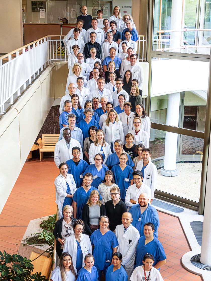 Group photo: Numerous people in white and blue medical uniforms are standing on a staircase, smiling. The photo was taken from an elevated perspective.