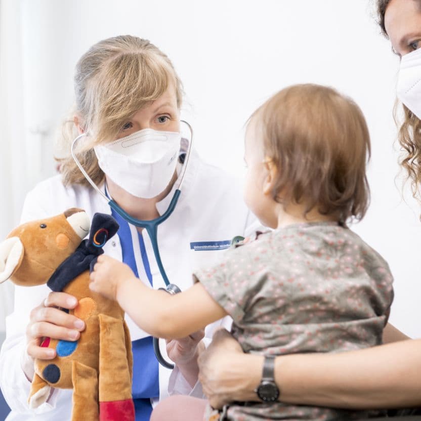 A doctor with a stethoscope and face mask sits opposite a small child. She is holding a cuddly toy deer, which the child is reaching for. The child is blurred in the background.
