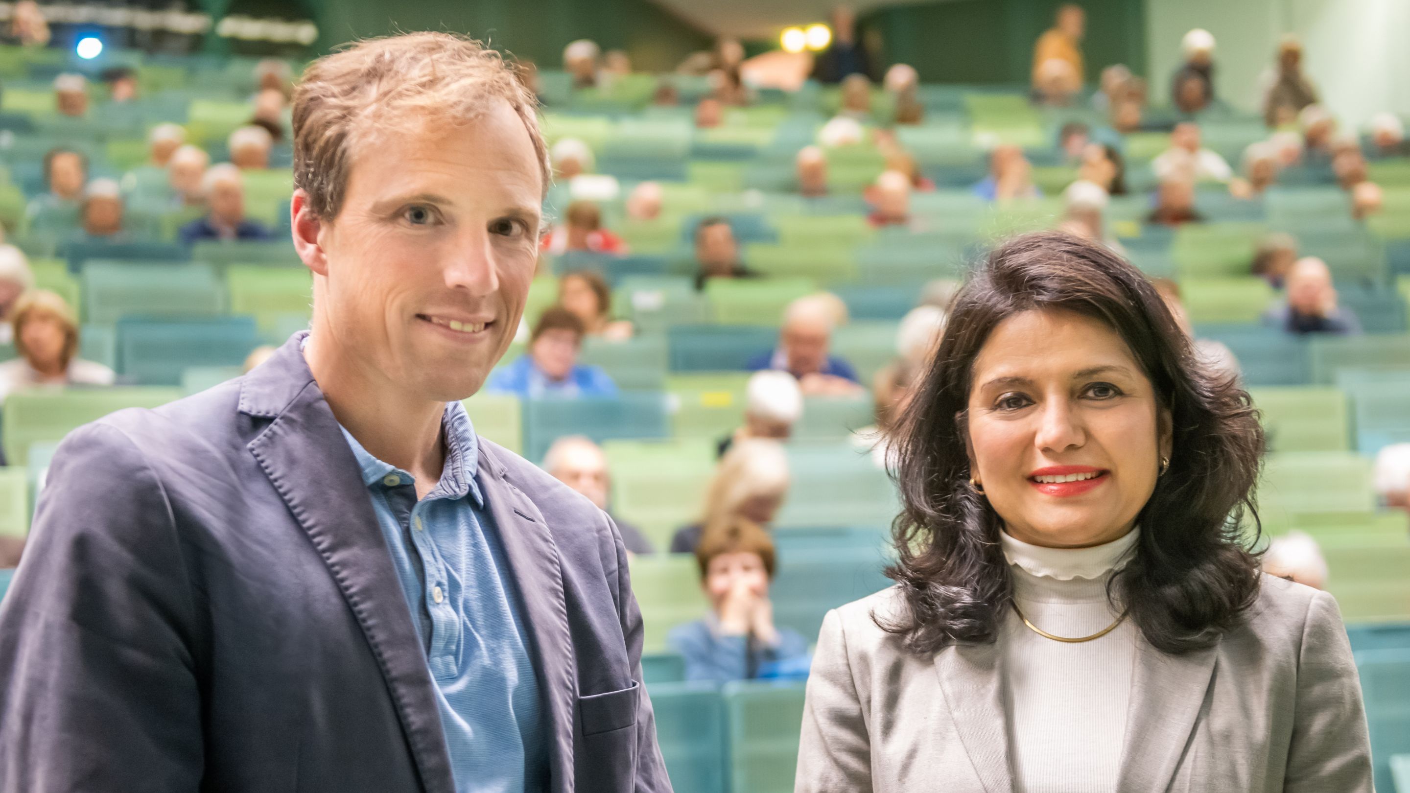 Prof. Dr. Jonas Tesarz and Prof. Dr. Rohini Kuner smile at the camera. The rows of a lecture hall can be seen blurred in the background.