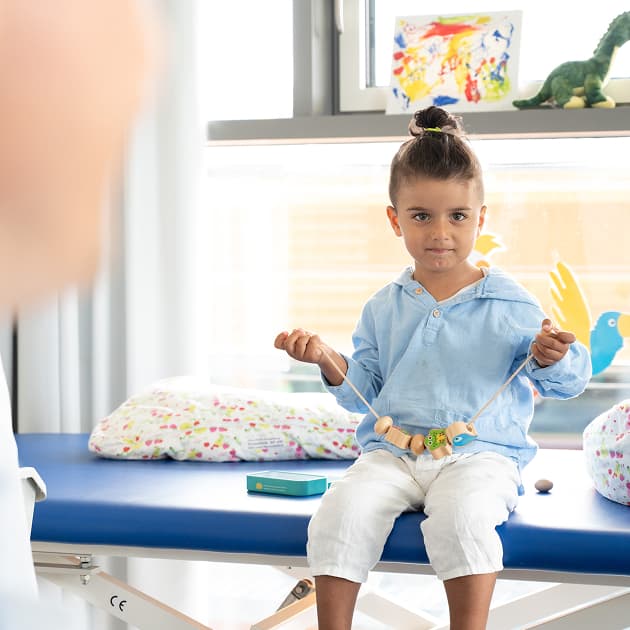 A child sits on a treatment table, holding a threading game in their hands and smiling softly at the camera. Colourful pictures hang in the background, with a stuffed animal beside them.