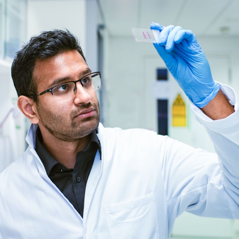 A person wearing a white coat and gloves holds a microscope slide in the air and looks at it.