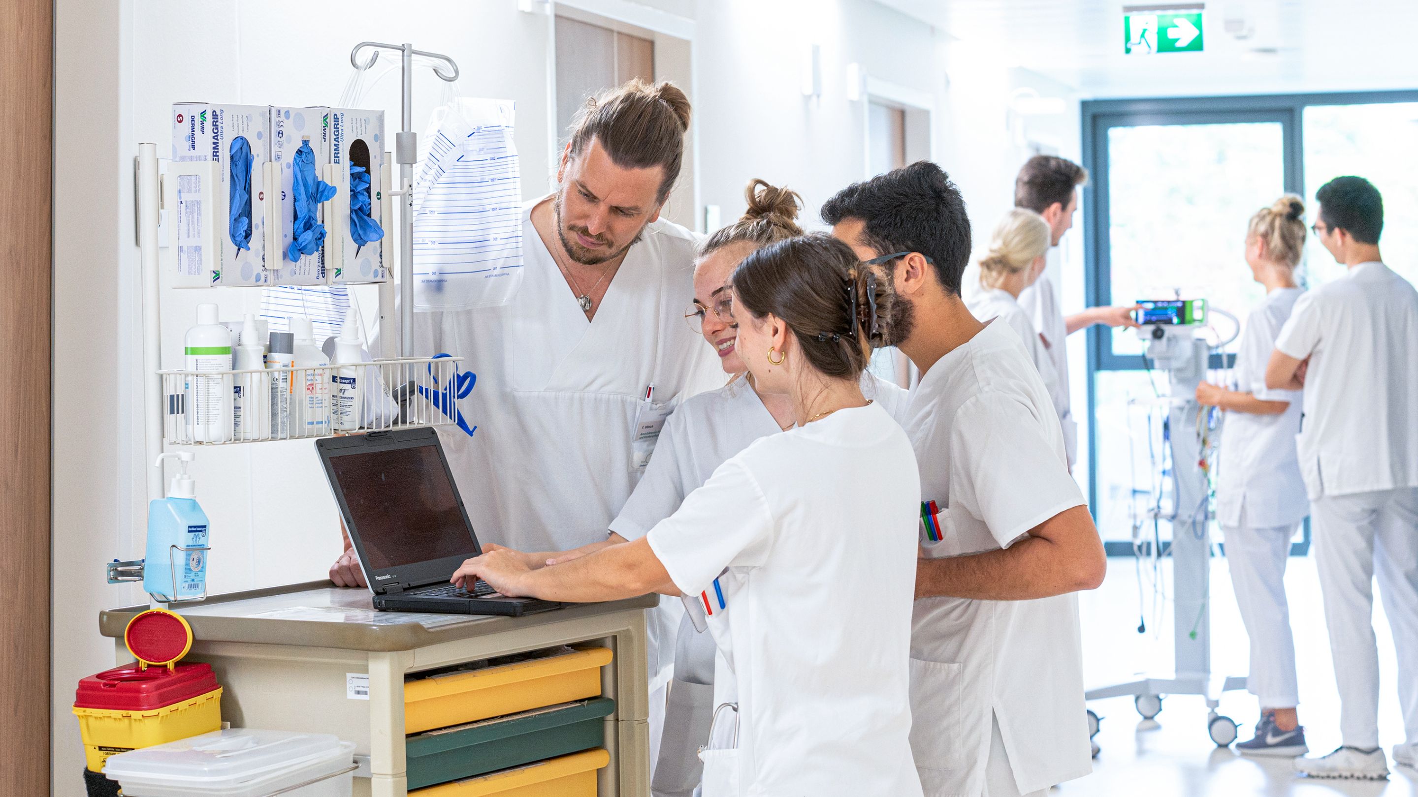 Ward corridor: In the foreground, a group of medical professionals stands around a trolley with a laptop. In the background, others in white coats can be seen, blurred, gathered around an IV stand.