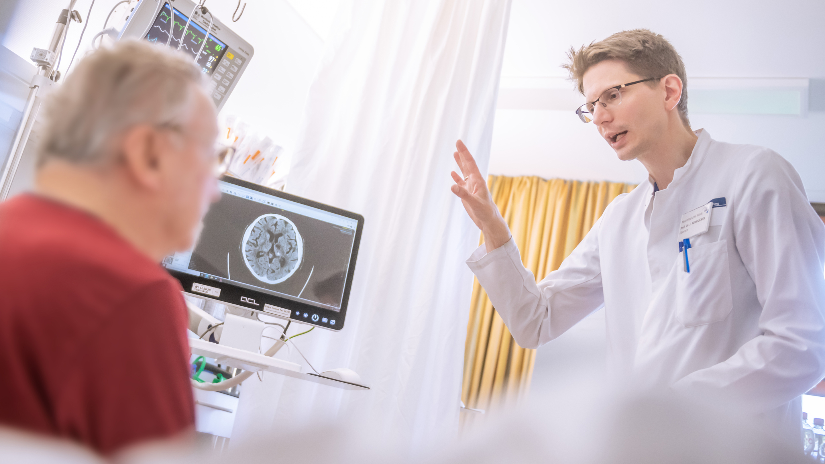 A medical professional in a white coat speaks animatedly to a patient. In the background, a monitor displays an MRI image of a head, with the patient blurred out in the background.