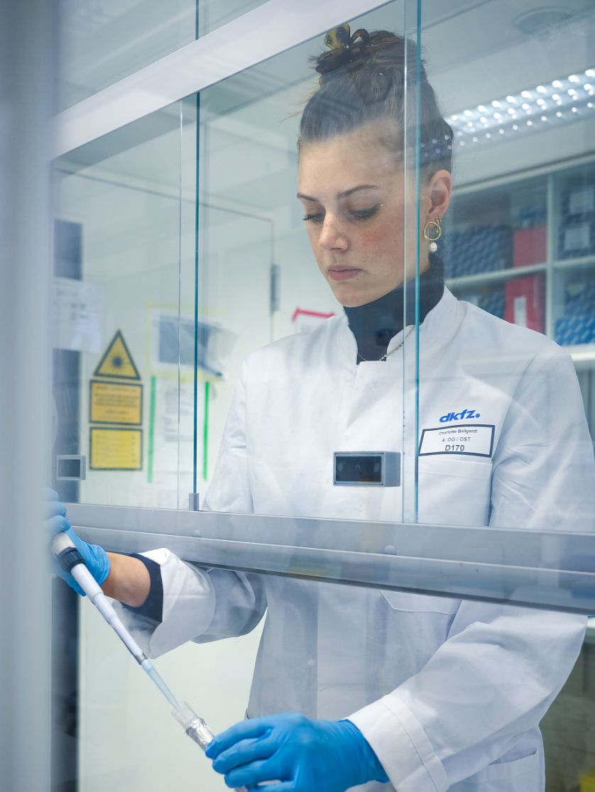 A person wearing lab clothing works with a pipette behind a protective glass screen.
