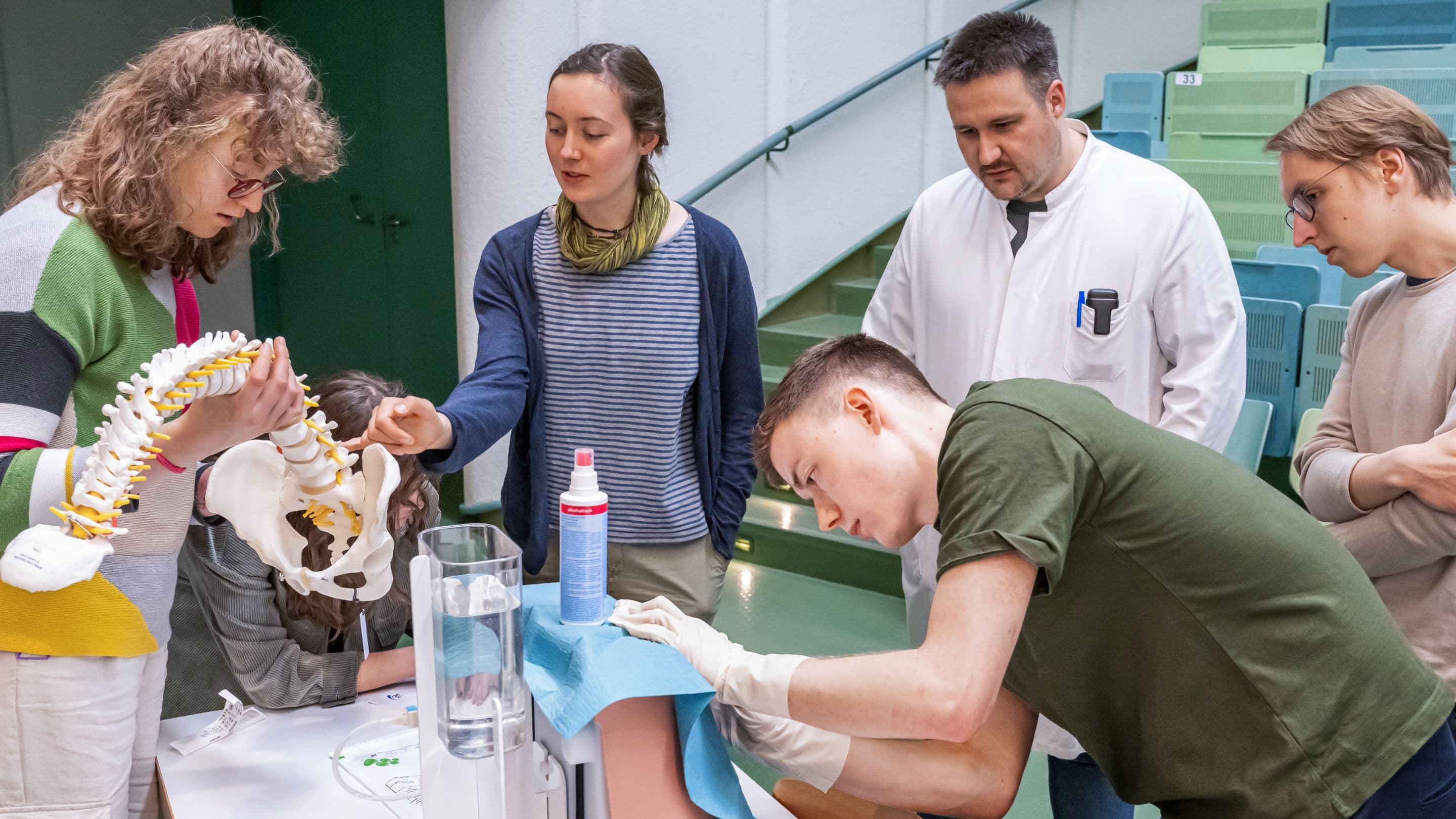 A group of students look at a model of the human spine together. In the foreground, a student leans over a back model with concentration. Next to him are a bottle of disinfectant and a medical professional in a white coat.