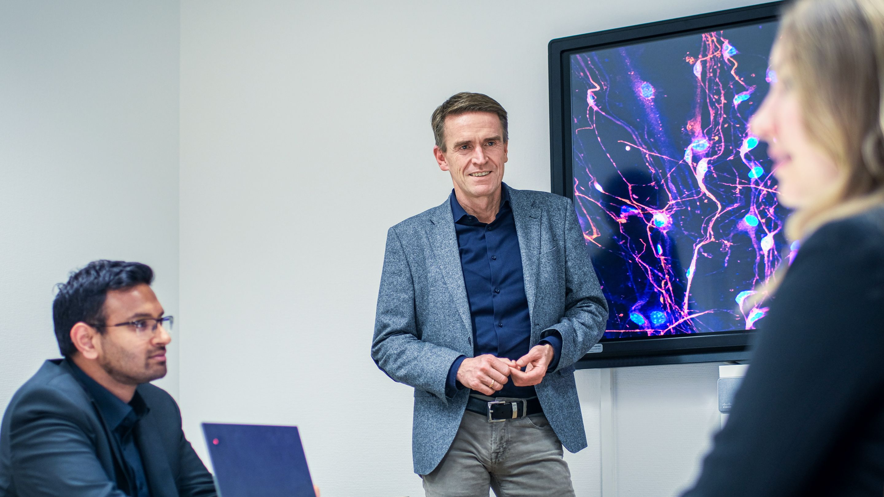 Prof. Dr. Wolfgang Wick with two other people in a meeting room. In the background, a large monitor displays bright lines and dots.