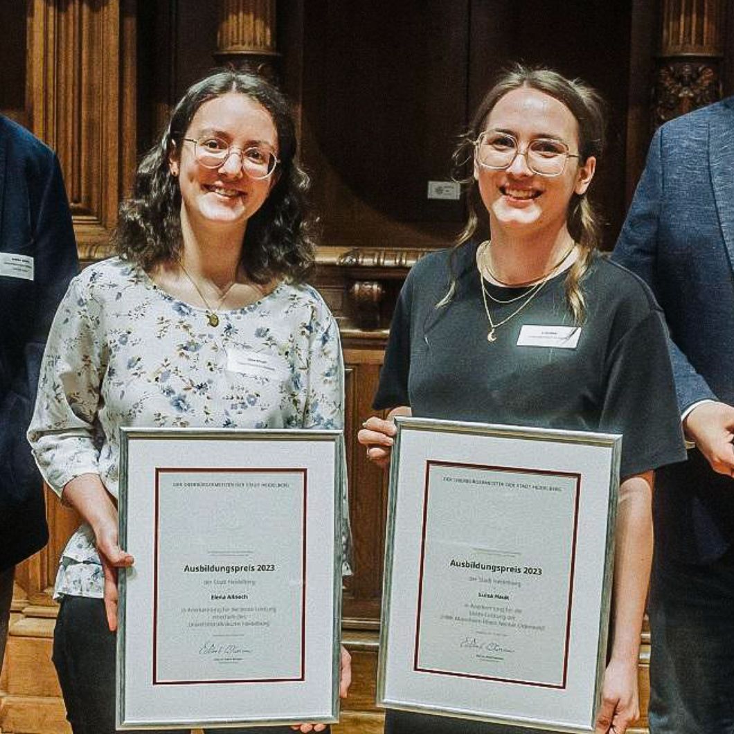 Group photo at the award ceremony: two female award winners with certificates in the middle, flanked by two men in suits. Everyone is smiling at the camera