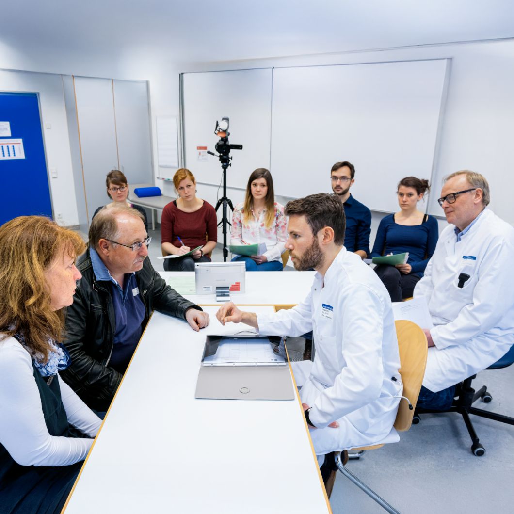 A medical professional is sitting at a table opposite two people, with a camera next to her for recording. In the background, there are other observers in everyday clothing and a doctor in a lab coat.