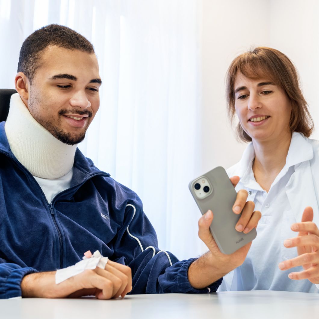 A patient wearing a neck brace sits at a table with a medical professional. He is holding a smartphone in his hand.