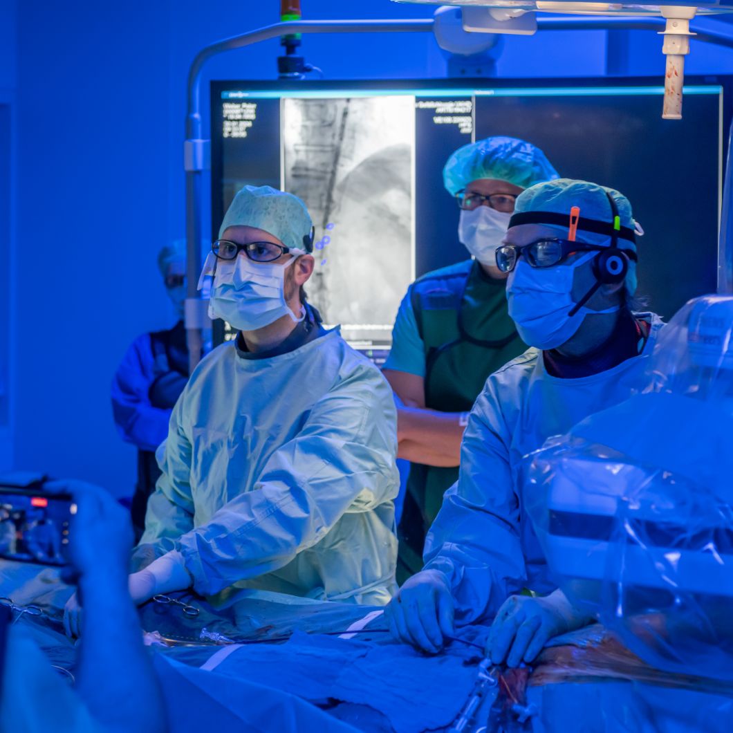 Three medical professionals in surgical attire performing an operation. The operating theatre is bathed in blue light, with a monitor displaying an X-ray image in the background.