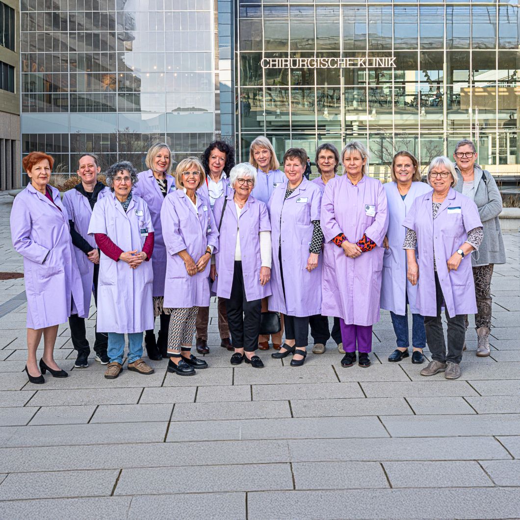 Group photo of the volunteer purple ladies and gentlemen in front of the entrance to the Medical and Surgical Clinic. The people are standing next to each other smiling in purple uniforms.
