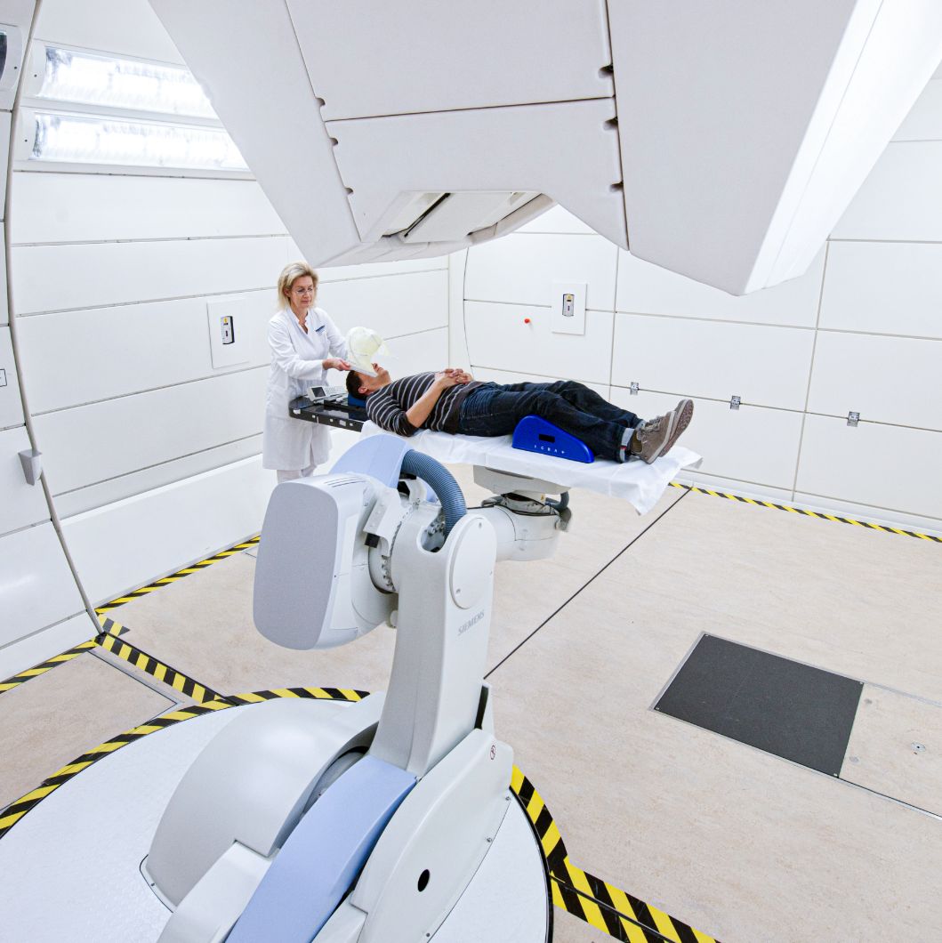 A patient lies in a treatment room at the Heidelberg Ion Beam Therapy Centre. A medical professional in a white coat stands at the head of the bed.