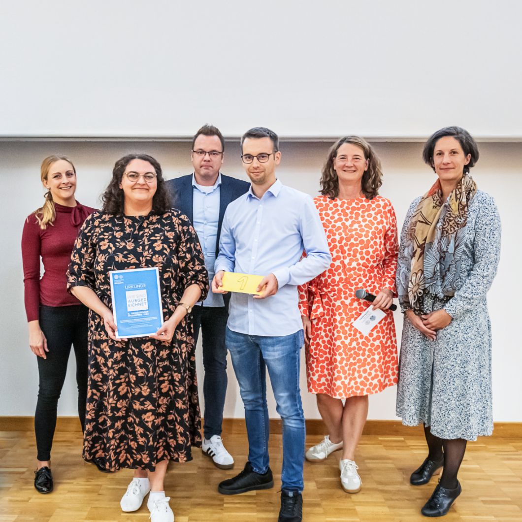 Group photo at the nursing award ceremony: Six people stand smiling next to a roll-up banner with the inscription &ldquo;Ausgezeichnet&rdquo; (Excellent).