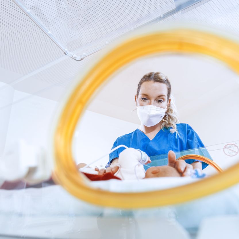 A nurse wearing a face mask leans over a premature baby in an incubator and gently places her hands on the child&rsquo;s head and stomach. The view is through the side opening of the incubator; the baby is blurred.