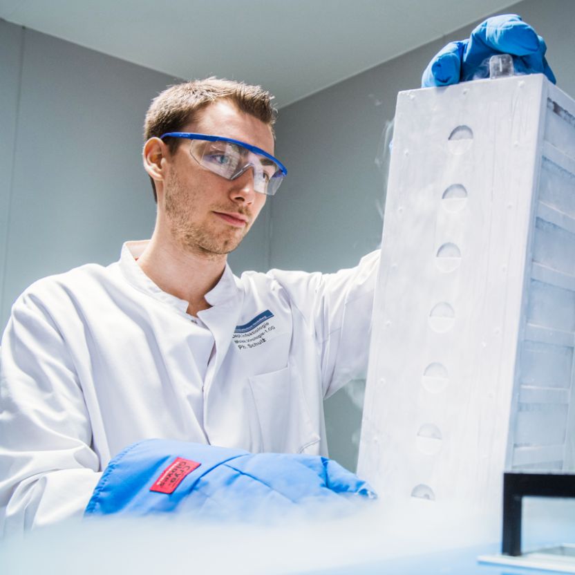 In a laboratory, a person wearing protective goggles and gloves lifts a frozen container. Cold mist rises visibly.