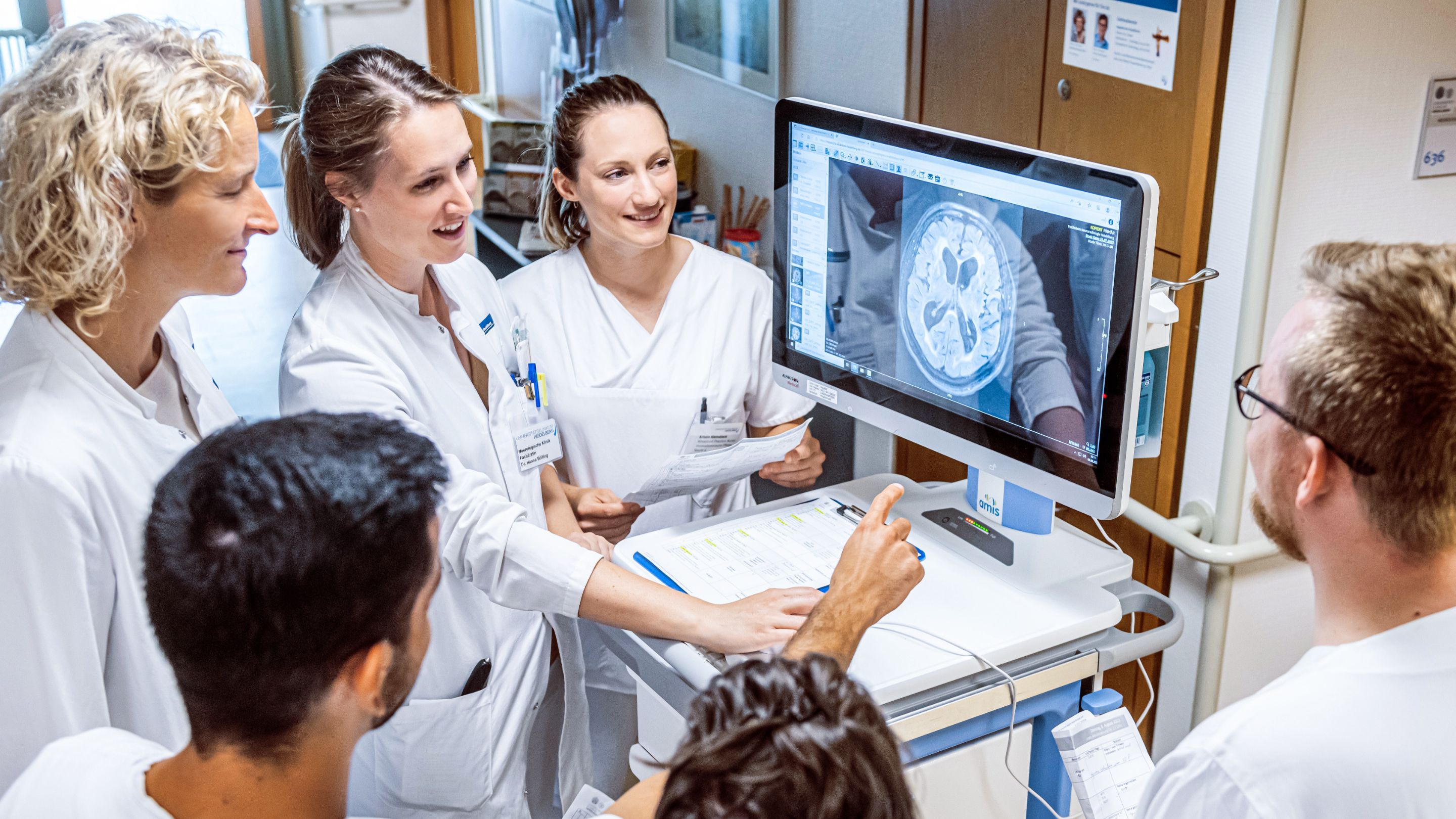 A small group of medical professionals look at an MRI scan of a head on a monitor. The screen is on a mobile trolley, with a ward corridor visible in the background.