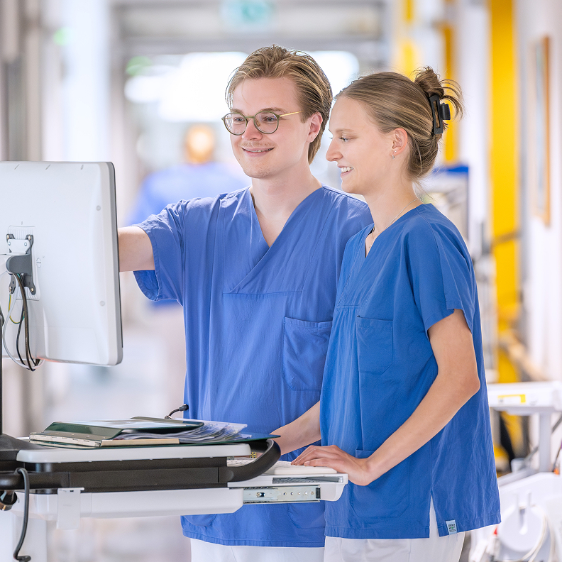 Two healthcare professionals in blue scrubs look at a computer screen together.