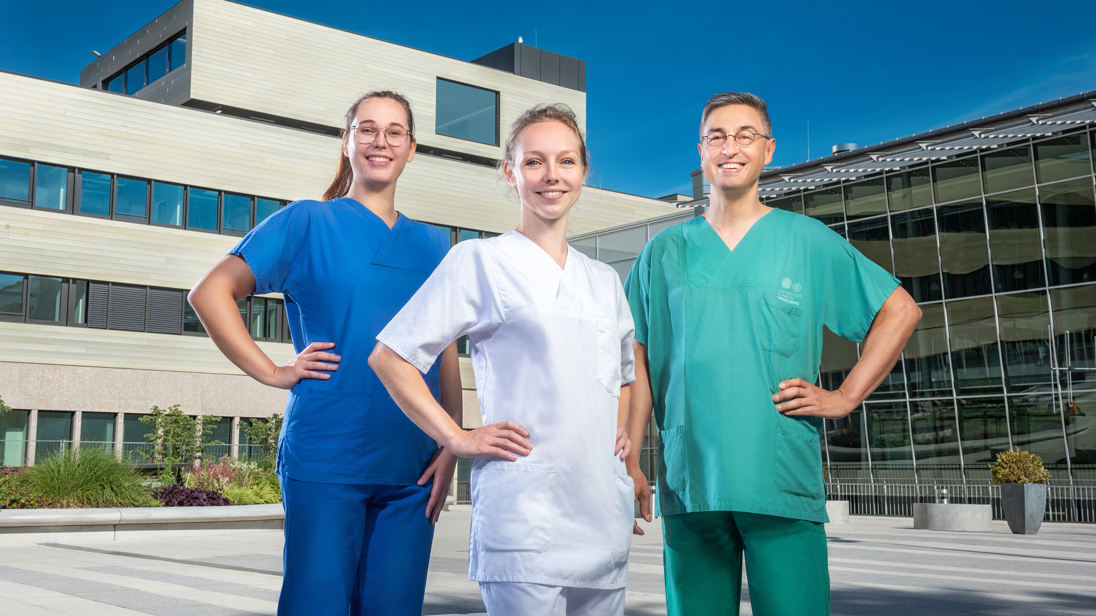 Three healthcare professionals in blue, white and green workwear stand in front of a hospital building.