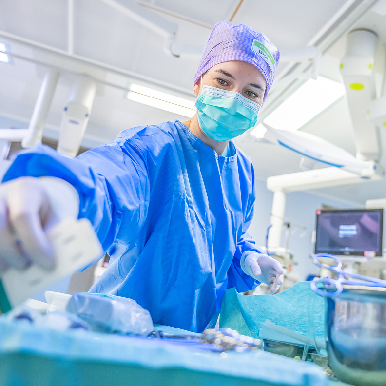 A healthcare professional wearing a face mask, surgical cap and blue protective gown prepares instruments for an operation.