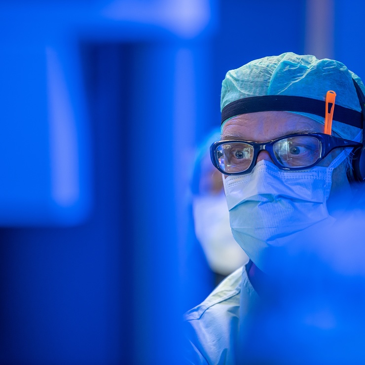 A healthcare professional wearing a face mask, protective goggles and surgical cap in a blue-lit environment.